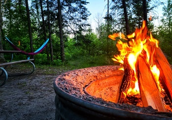 A roaring bonfire in a rustic campfire ring, an empty hammock sways in the trees beyond as the sun sets behind the forest.