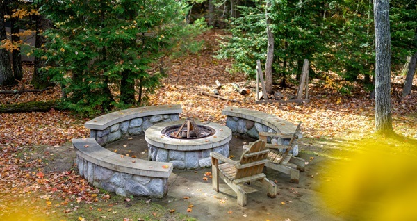 A stone fire pit sits serenely amongst a sunlit forest, two adarondack chairs beckoning invitingly.