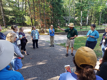 A group of people stand around a presenter wearing DNR green as they gesture to the forest around them.