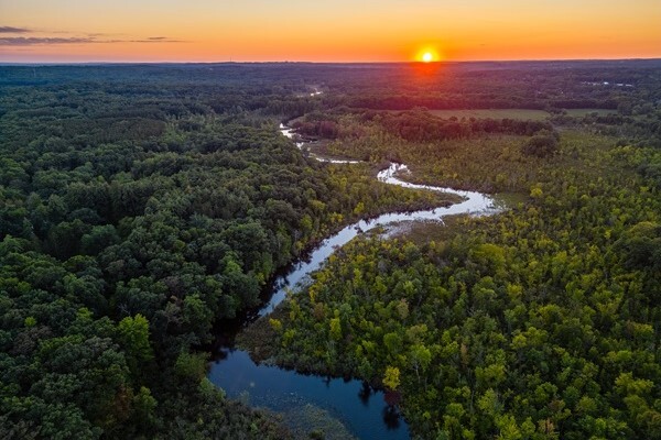 A sunset illuminates Proud Lake, casting the water in a glassy glow amidst a dark, green forest.