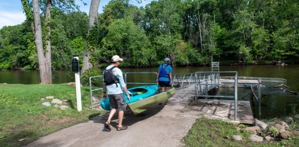 Two adults carry a kayak down a paved trail to an accessible kayak launch on a river lined with trees at Delta Mills Park in Lansing, Michigan.