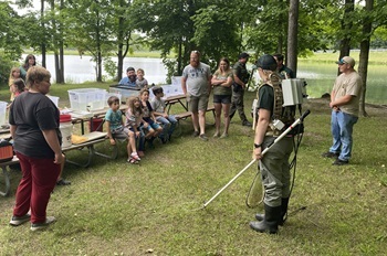 a woman in T-shirt and waders holds electrofishing gear while talking to kids and adults sitting at picnic tables outdoors