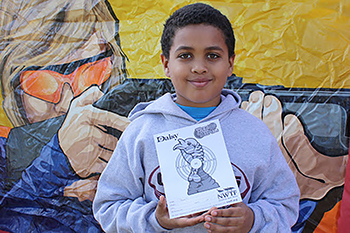 a smiling, dark-haired boy in a gray sweatshirt holds a paper with an illustrated turkey and target. He is standing in front of a colorful banner