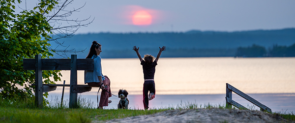 a mother sitting on a bench watching her child jump up facing lake with sunset