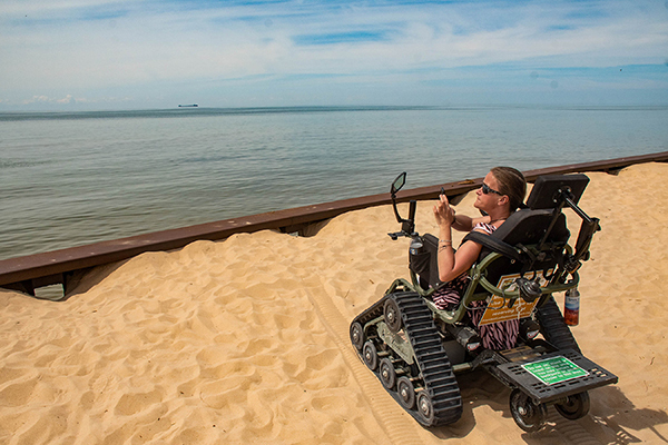 A woman sits in a track chair taking a picture with her phone on a sandy beach looking out into Lake Michigan.