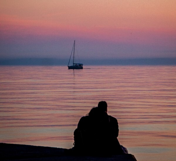 A couple watch a lone sailboat from the pier at sunset.