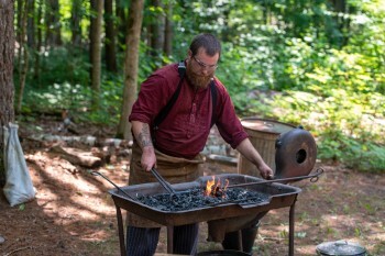 A blacksmith hammers a red-hot rod at an outdoor forge. 