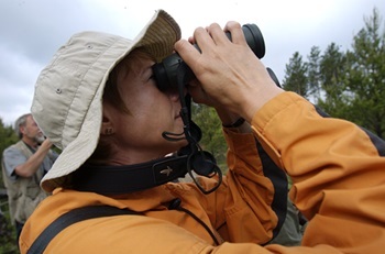 short-haired woman in tan, floppy hat and orange jacket looks through black binoculars