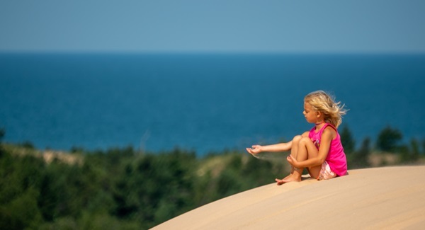 blond little girl in a pink shirt and shorts sits on a sand dune, sand streaming from her hand, against bright blue sky and Lake Michigan