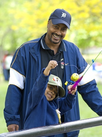 Man in a navy blue jacket and Detroit Tigers ballcap and a little girl in a ballcap both smile as she shows off her caught fish and pole.