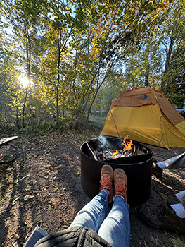 person with feet up near campfire on wooded campsite in morning