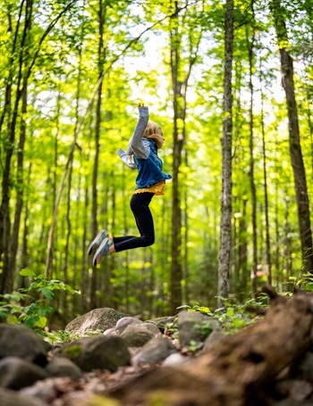 young girl in black leggings, sneakers and jacket stretches arms up while jumping over rocks, logs and grass in forest at Bewabic State Park