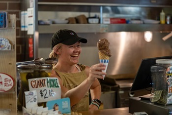 a smiling young woman in a T-shirt, apron and baseball cap hands a chocolate ice cream cone across a counter with a cash register and pickle jar