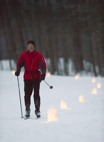 a man in a black cap, red winter jacket and black ski pants uses poles to cross-country ski on a snow-covered, lantern-lit trail in the woods