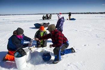 A small group of adults and kids in winter outdoor wear have ice-fishing gear around a hole in the ice of a frozen lake at Mitchell State Park