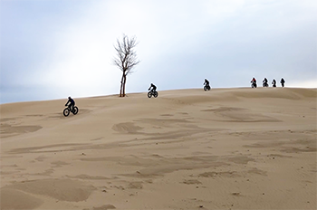 several fat-tire bikers riding over a sandy dune all bundled up