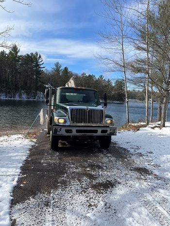 front view of a silver and black fish stocking truck, with its back end at the end of an icy drive leading into a large, tree-lined body of water