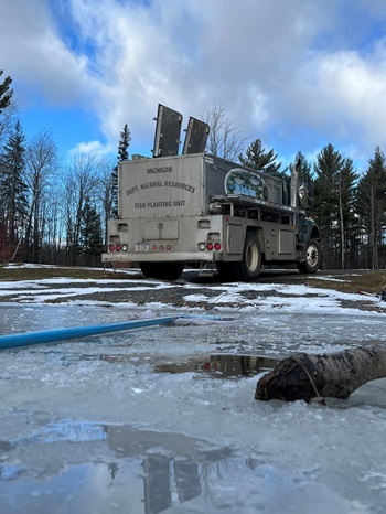 The back end of a silver fish-stocking truck, imprinted with the words DNR Fish Planting Unit, on the bank of an icy body of water in forested area