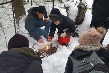 A small group of adults in winter gear huddle around a small campfire in the snow, as one of them works to stoke the flames