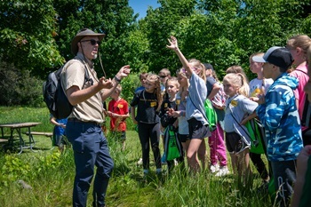 a man in blue jeans, tan shirt and brimmed hat talks with a dozen children outdoors at Sleepy Hollow State Park on a Nature Awaits tour