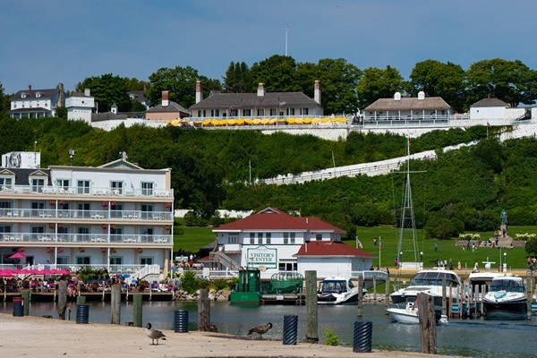 several boats in slips next to a visitors center and a multistory condo along Mackinac Island State Harbor, with houses on the rising bluffs