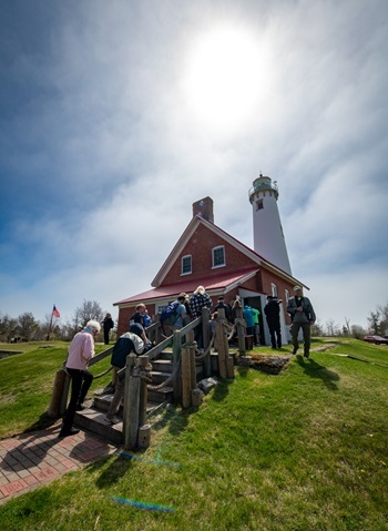 people line up on the stairs up to the newly restored, red and white Tawas Point Lighthouse, with bright sun overhead