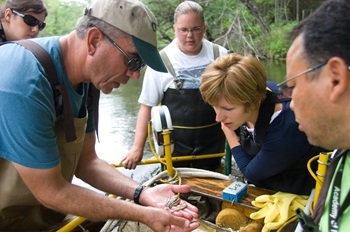a man in waders holds a small fish in his hands as two women and another man look on. They're along the Au Sable River.