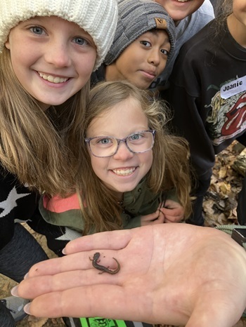 a couple girls and boys in hats and jackets smile while looking at a salamander stretched out in an adult's hands