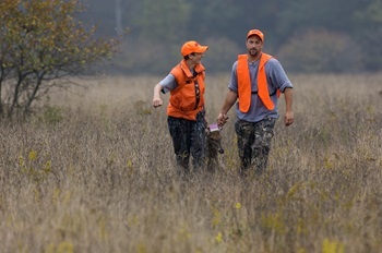 a teen boy and a young man, both wearing hunter orange caps and vests, pull a tagged buck through tall grass
