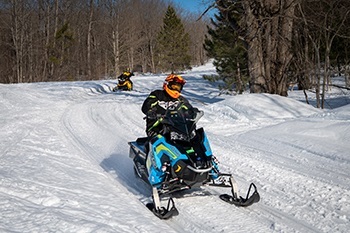 person in orange helmet and winter gear on a black and blue snowmobile stops on a curve of a groomed, snowy trail in a Michigan forest