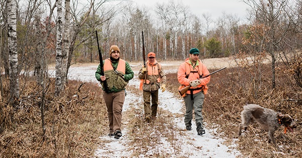 three hunters in camo and hunter orange clothing carry guns while walking through a snowy forest, with a dog running slightly ahead