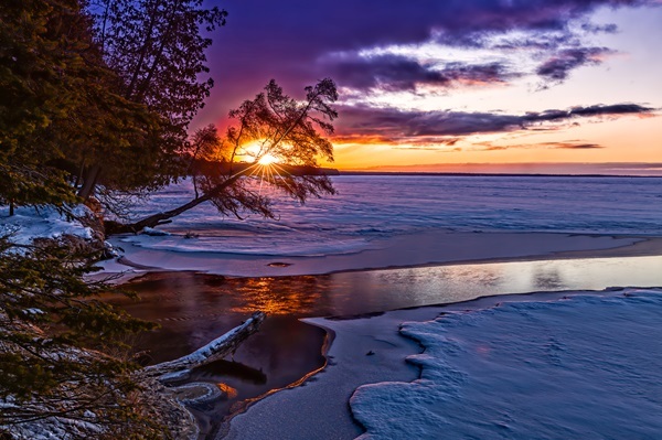 Bright orange sunrise breaks through a blue-purple sky and tree over North Higgins Lake, with ice and snow covering the near shore
