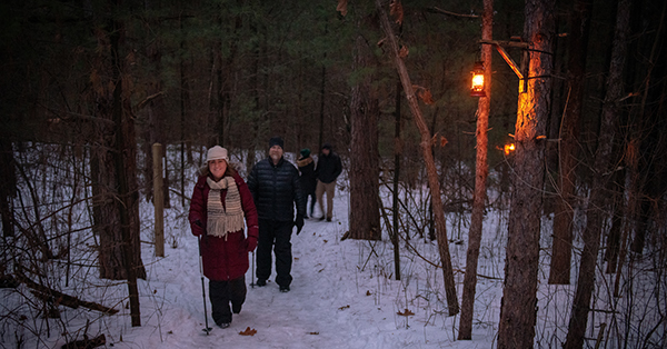 people hiking on snowy, wooded trail lit by lantern