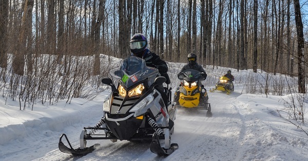 snowmobiles moving down a sunlit, groomed, snow-covered trail in a forest in Gogebic County, Michigan.