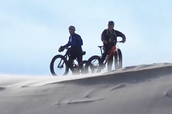 two adults in helmets standing with their fat-tire bikes, nearly silhouetted atop a large sand dune, against a cold, pale blue sky