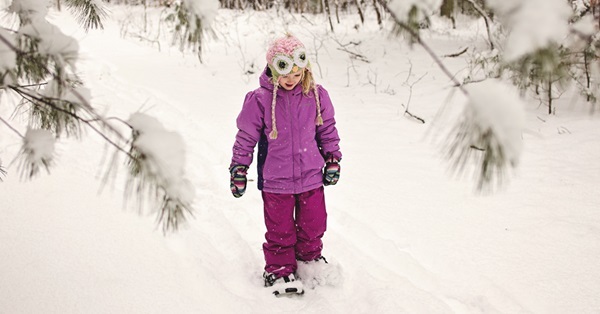 smiling little girl in purple jacket and snow pants and a pink hat with google eyes walks through a snowy forest trail