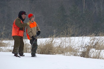Older man and younger boy, both in camo and hunter orange gear, stand in a snowy field looking ahead