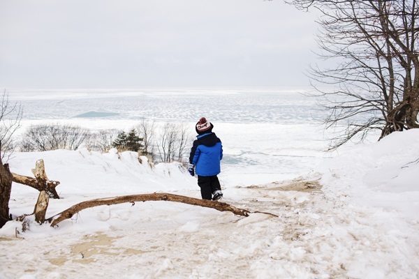 young child dressed in blue and black snow coat, pants and knit hat stands on a snowy shore, looking out at icy Lake Michigan