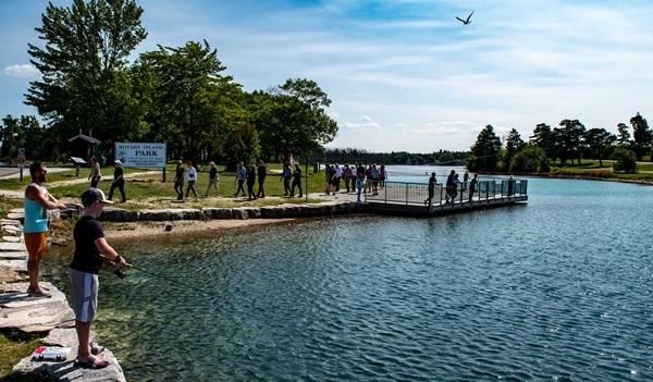 Dozens of kids and adults stroll on and fish from a pier and stone-lined shoreline in Rotary Island Park, Sault Ste. Marie, Michigan