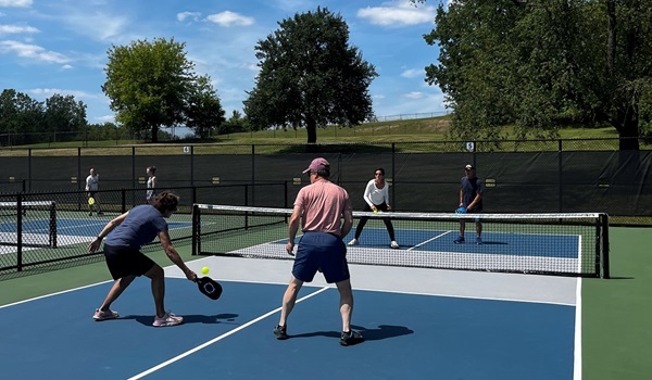 Men and women in summer clothing, using racquets, playing pickleball on a new, green-and-blue court set in a forested park area