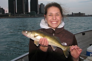 a young girl in a vest and hoodie, smiling broadly, shows off a walleye caught on the Detroit River; city buildings in the background