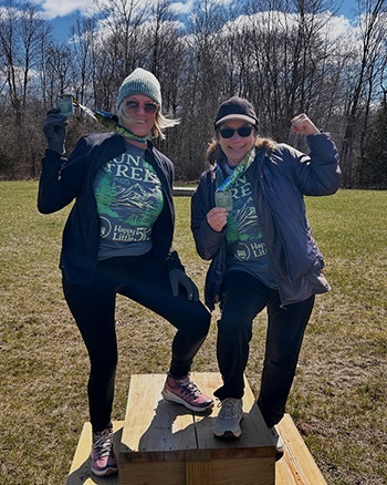 two women in running leggings, race T-shirts and finisher medals celebrate participating on a Happy Little 5K race