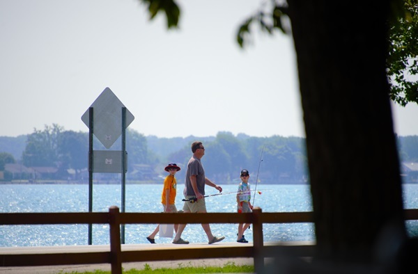 Father and two young sons in summer clothing carry fishing poles and gear on a shoreline walkway at Algonac State Park