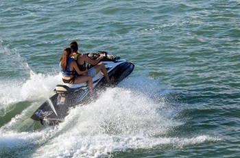 two people in bathing suits and life vests right on a personal watercraft, leaving a foamy wake in deep-blue water