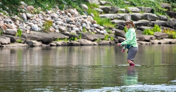 woman in shorts, jacket and hat holds her fly fishing rod low over a calm area of Battle Creek