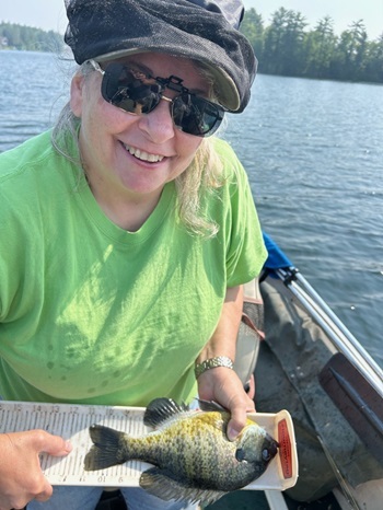smiling woman in green T-shirt, sunglasses and hat sits in a boat on water, holding a bluegill on a long, white measuring board