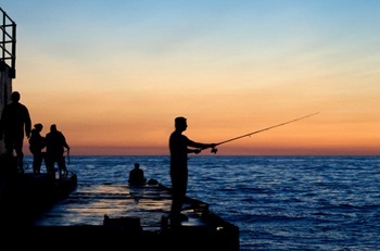 man fishing off a pier, with other people in shadow, set against an orange sky and large blue body of water