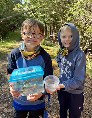 two smiling, young boys, one in glasses and holding a frog in tank, the other in a hoodie and holding a cup, in the woods on a sunny day