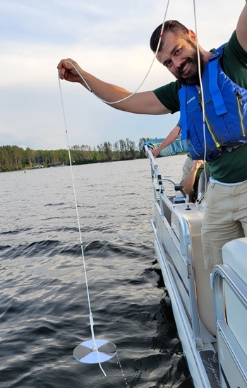 A Cooperative Lakes Monitoring Program volunteer on a boat collects water clarity measurements with a Secchi disk hanging in water