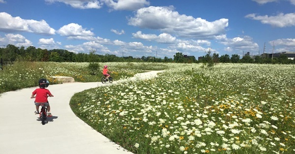 two little kids wearing helmets and summer clothes ride bikes on a winding, paved path amid grassy, flowered grounds